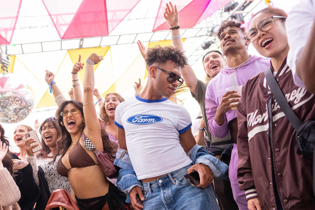 A group of young people are dancing and cheering under colorful triangular canopy lights at an outdoor event, looking joyful and energetic.